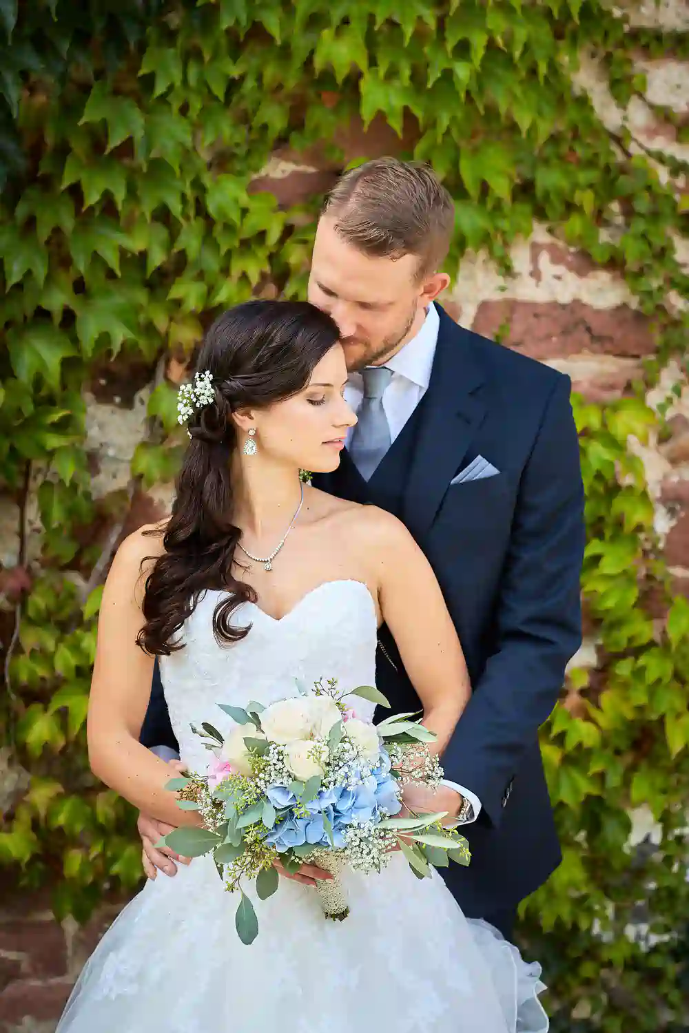 Onelovephoto Eine Braut in einem weißen Kleid mit einem Blumenstrauß in der Hand steht vor einem Bräutigam in einem dunklen Anzug, der sie sanft von hinten umarmt, vor einer efeubewachsenen Backsteinmauer - aufgenommen als Teil einer Hochzeitsreportage im Hotel Grenzhof Heidelberg. Hochzeitsfotografie in Mannheim und Heidelberg