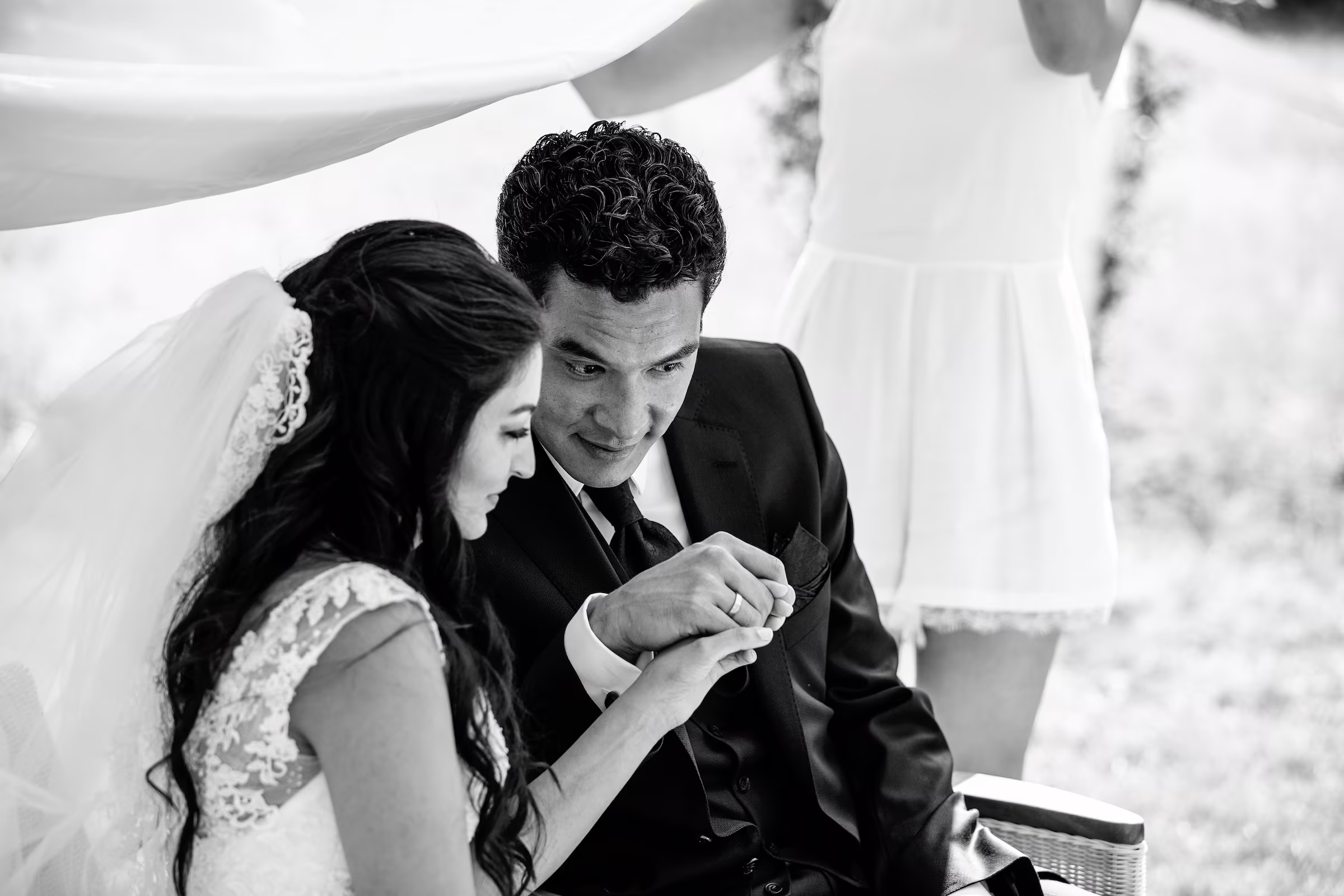 Eine Braut und ein Bräutigam sitzen eng beieinander, halten sich an den Händen und lächeln, während eine Frau in einem kurzen weißen Kleid während einer Hochzeitszeremonie im Freien ein Tuch über sie hält. Das Foto wurde von einem Hochzeitsfotografen in der Nähe von Mannheim aufgenommen und ist in schwarz-weiß gehalten.