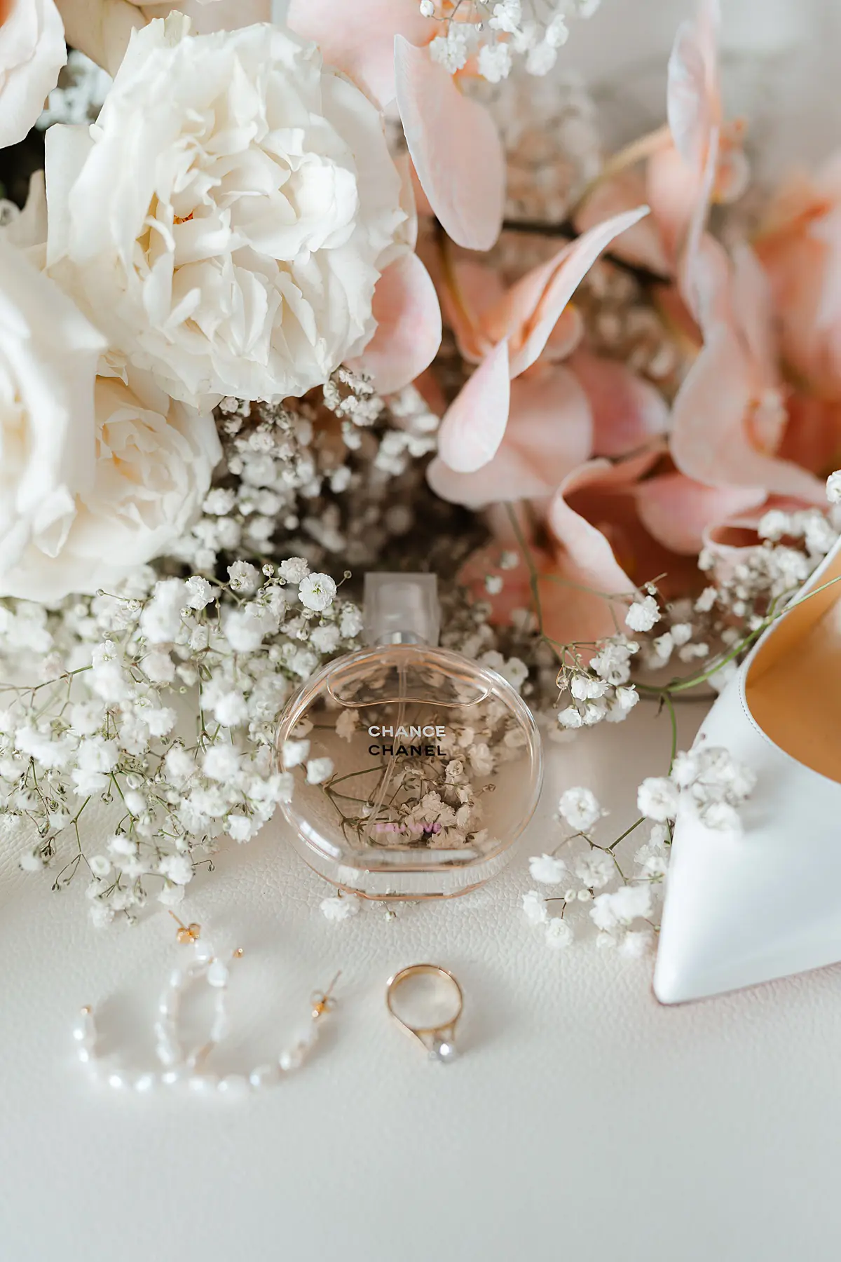 Onelovephoto Ein Flakon des Parfums Chanel Chance steht auf einer weißen Fläche, umgeben von weißen und rosafarbenen Blumen, einem Perlenohrring, einem goldenen Ring mit einer Perle und der Spitze eines weißen Schuhs. Hochzeitsfotografie in Mannheim und Heidelberg