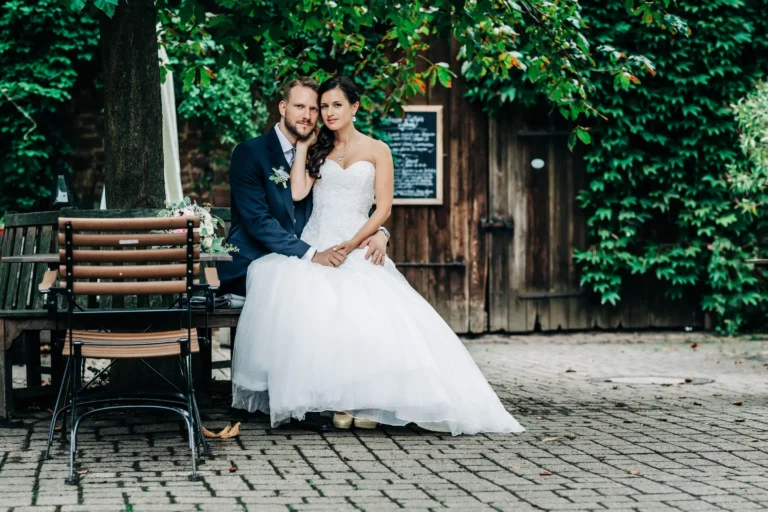 Eine Braut in einem weißen Kleid und ein Bräutigam in einem marineblauen Anzug sitzen zusammen auf einer Bank im Freien des Hotels Grenzhof Heidelberg, umgeben von Grün und rustikalen Holzgebäuden - ein perfekter Moment für ihre Hochzeitsreportage, entspannt und glücklich wirkend.