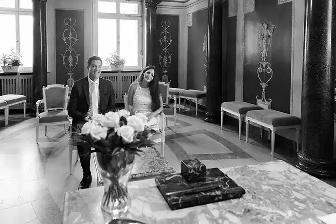 Ein lächelndes Paar sitzt Seite an Seite in einem formellen Raum mit kunstvollem Dekor und großen Fenstern. Als Hochzeitsfotograf in Heidelberg und Mannheim fange ich zeitlose Schwarz-Weiß-Momente wie diesen ein, mit zarten Blumen auf einem Marmortisch im Vordergrund. Onelovephoto - Hochzeitsfotografie in Mannheim und Heidelberg