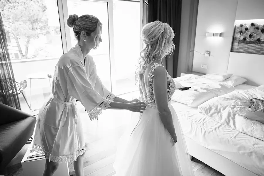 Onelovephoto Eine Frau im Bademantel hilft einer Braut in einem hellen, modernen Schlafzimmer in ihr Hochzeitskleid. Die Szene ist offen, mit ungemachten Betten und natürlichem Licht, das durch große Fenster strömt. Hochzeitsfotografie in Mannheim und Heidelberg