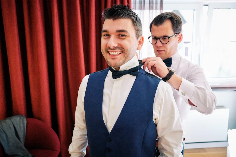 Onelovephoto Ein lächelnder Mann in einer blauen Weste steht, während ein anderer Mann ihm hilft, seine Fliege zu richten. Sie befinden sich in einem Innenraum, in der Nähe eines Fensters mit roten Vorhängen im Hintergrund. Hochzeitsfotografie in Mannheim und Heidelberg
