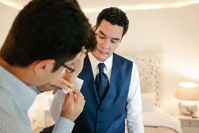 Onelovephoto Ein Mann in blauer Weste und Krawatte steht in einem Schlafzimmer, während ein anderer Mann ihm hilft, seinen Hemdsärmel zu richten. Der Raum ist sanft beleuchtet, im Hintergrund sind eine Lampe und ein Bett zu sehen. Hochzeitsfotografie in Mannheim und Heidelberg