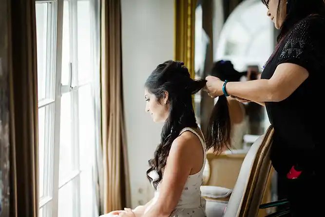 Eine Frau sitzt am Fenster und schaut nach draußen, während eine andere Person ihr langes dunkles Haar zu einer eleganten Hochsteckfrisur frisiert. Als Hochzeitsfotograf in Heidelberg und Mannheim halte ich solche natürlichen, lichtdurchfluteten Momente für euch fest. Onelovephoto - Hochzeitsfotografie in Mannheim und Heidelberg