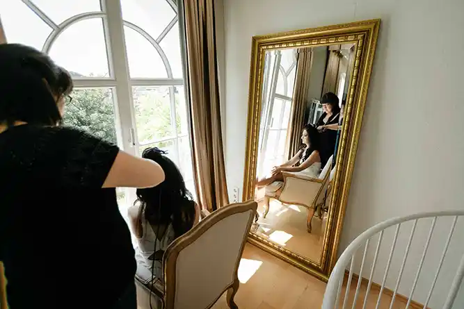 Onelovephoto Eine Frau stylt einer anderen Frau in der Nähe eines großen Fensters die Haare; beide spiegeln sich in einem hohen, verzierten Spiegel, wobei natürliches Licht den eleganten Raum erfüllt. Hochzeitsfotografie in Mannheim und Heidelberg