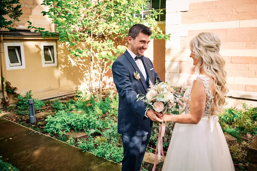Onelovephoto Ein Bräutigam in einem marineblauen Anzug und mit Fliege lächelt, während er mit einer Braut in einem weißen Kleid und mit einem Blumenstrauß in der Hand in einem sonnenbeschienenen Garten in der Nähe einer Mauer steht. Hochzeitsfotografie in Mannheim und Heidelberg