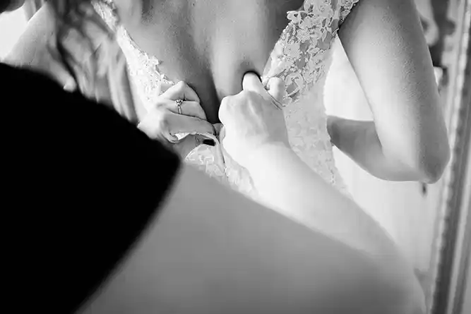 Onelovephoto Schwarz-Weiß-Foto einer Person in Nahaufnahme, die einer Braut ein Hochzeitskleid aus Spitze anlegt, wobei der Schwerpunkt auf ihren Händen und dem zarten Stoff liegt. Hochzeitsfotografie in Mannheim und Heidelberg