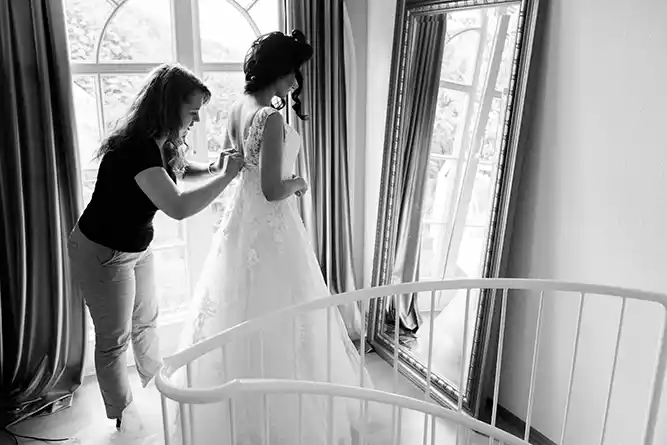 Onelovephoto Eine Frau hilft einer Braut beim Anpassen ihres Kleides in der Nähe eines großen Fensters und eines Ganzkörperspiegels, durch den weiches Licht fällt. Die Braut trägt ein weißes Kleid, und die Szene ist in Schwarz-Weiß gehalten. Hochzeitsfotografie in Mannheim und Heidelberg