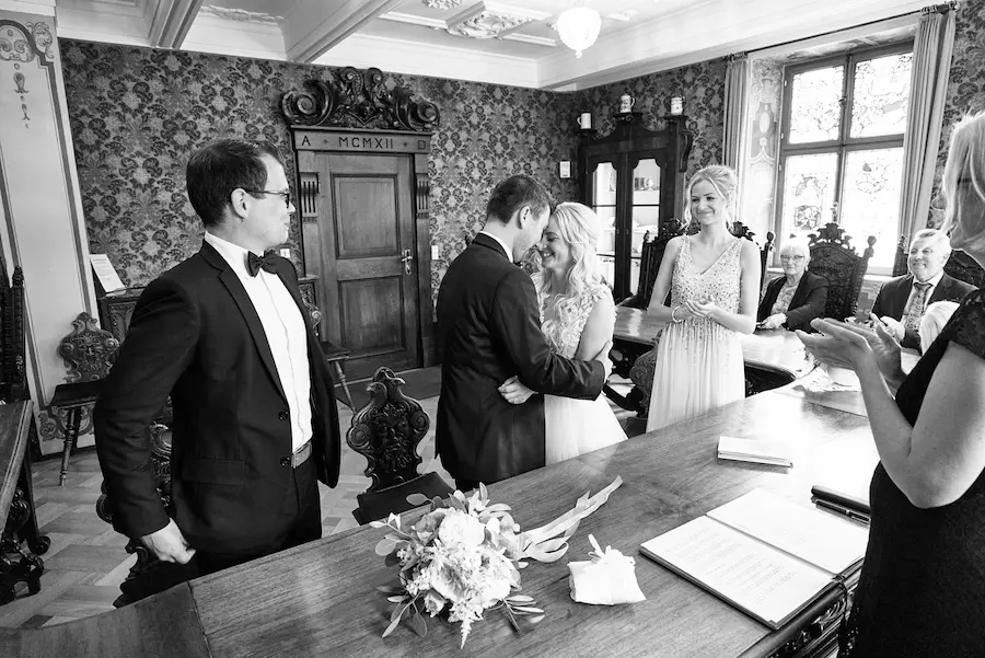 Eine Braut und ein Bräutigam umarmen sich und lächeln am Altar während einer Hochzeitszeremonie in einem geschmückten Raum. Die Gäste applaudieren, als Jessie und Marcel JA - Heiraten in Deidesheim sagen, mit einem Blumenstrauß und einem offenen Buch auf dem Tisch im Vordergrund. Onelovephoto - Hochzeitsfotografie in Mannheim und Heidelberg