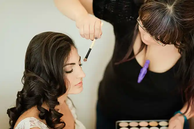 Onelovephoto Eine Maskenbildnerin trägt Lidschatten auf eine Frau mit langem, gelocktem Haar auf, die mit geschlossenen Augen dasitzt. Die Künstlerin hält eine Palette und einen Pinsel in der Hand und konzentriert sich auf das Gesicht der Frau. Hochzeitsfotografie in Mannheim und Heidelberg
