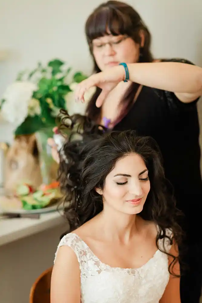Onelovephoto Eine Braut mit langen dunklen Haaren und einem Spitzenkleid sitzt gelassen da, während eine Friseurin ihr Haar stylt, mit Blumen und Schönheitswerkzeugen auf dem Tresen im Hintergrund. Hochzeitsfotografie in Mannheim und Heidelberg