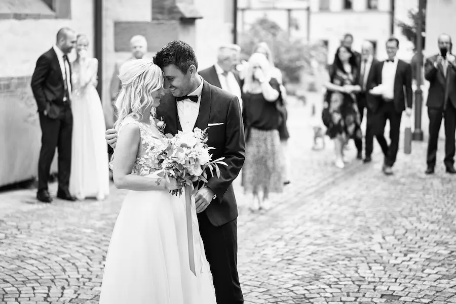 Onelovephoto Eine Braut und ein Bräutigam umarmen sich und lächeln sich auf einer Kopfsteinpflasterstraße an, umgeben von Freunden und Familie in formeller Kleidung. Das Bild ist in schwarz-weiß gehalten. Hochzeitsfotografie in Mannheim und Heidelberg