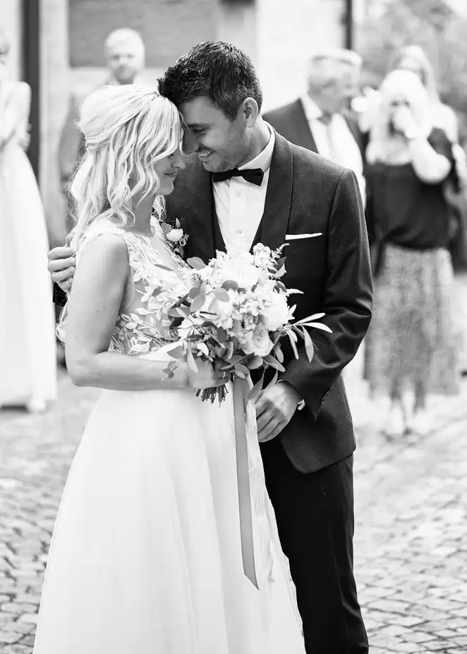 Braut und Bräutigam, Jessie und Marcel, stehen eng beieinander, lächeln und berühren sich an der Stirn. Das Bild fängt den Moment ihrer Hochzeit in Deidesheim in Schwarz-Weiß ein, wobei einige Gäste im Hintergrund verschwommen sind. Braut und Bräutigam, Jessie und Marcel, stehen eng beieinander, lächeln und berühren sich an der Stirn. Das Bild fängt den Moment ihrer Hochzeit in Deidesheim in Schwarz-Weiß ein, wobei einige Gäste im Hintergrund verschwommen sind.