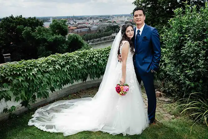 Onelovephoto Eine Braut in einem weißen Kleid und Schleier steht neben einem Bräutigam in einem blauen Anzug, der einen bunten Blumenstrauß hält. Sie posieren im Freien vor einer grünen Kulisse, mit einer Stadtlandschaft und einem Fluss im Hintergrund. Hochzeitsfotografie in Mannheim und Heidelberg