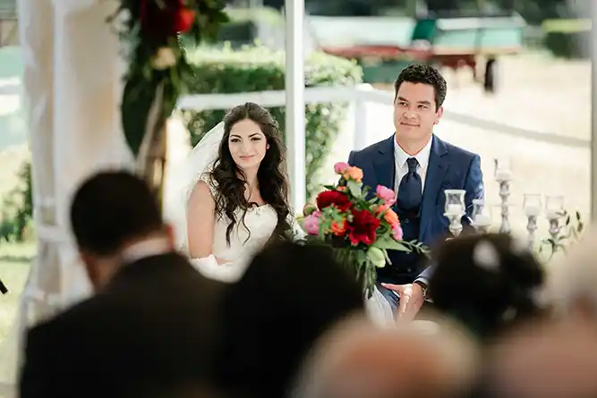 Onelovephoto Eine Braut in einem weißen Kleid und ein Bräutigam in einem blauen Anzug sitzen Seite an Seite und lächeln während einer Hochzeitszeremonie, mit bunten Blumen auf dem Tisch vor ihnen und Gästen im Vordergrund. Hochzeitsfotografie in Mannheim und Heidelberg