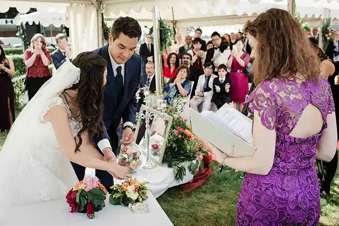 Onelovephoto Eine Braut und ein Bräutigam nehmen an einer Hochzeitszeremonie im Freien unter einem weißen Zelt teil, umgeben von Gästen. Eine Frau in einem lilafarbenen Spitzenkleid nimmt die Trauung vor und hält eine Mappe in der Hand, während das Paar mit bunten Blumen auf einem Tisch spielt. Hochzeitsfotografie in Mannheim und Heidelberg