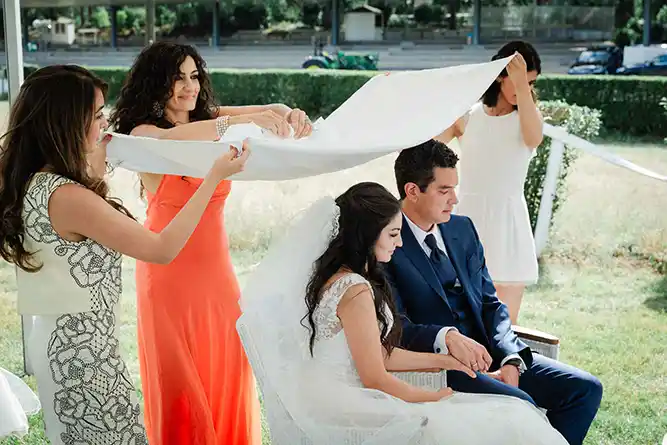 Onelovephoto Braut und Bräutigam sitzen bei einer Hochzeitszeremonie im Freien nebeneinander, während zwei Frauen ein weißes Tuch über ihre Köpfe halten und eine weitere Frau zusieht. Hochzeitsfotografie in Mannheim und Heidelberg