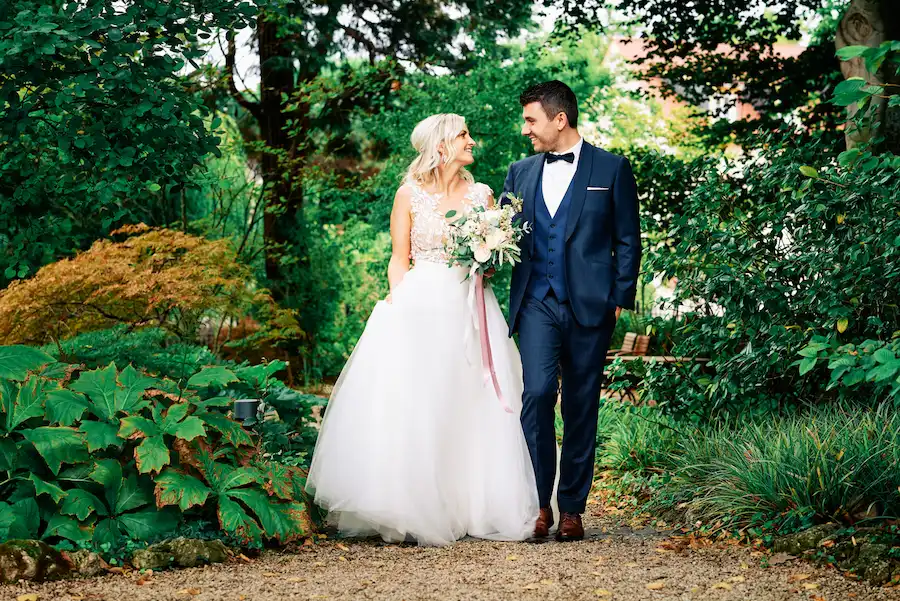 Onelovephoto Eine Braut in einem weißen Kleid und ein Bräutigam in einem dunklen Anzug gehen zusammen auf einem Gartenweg, lächeln sich an und sind von üppigem Grün umgeben. Hochzeitsfotografie in Mannheim und Heidelberg