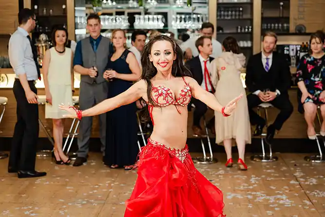 Onelovephoto Eine Frau in einem roten Bauchtanzkostüm tritt vor einer Gruppe von Menschen in einer Bar auf, die ihr interessiert zusehen. Einige sitzen, während andere im Hintergrund stehen. Hochzeitsfotografie in Mannheim und Heidelberg