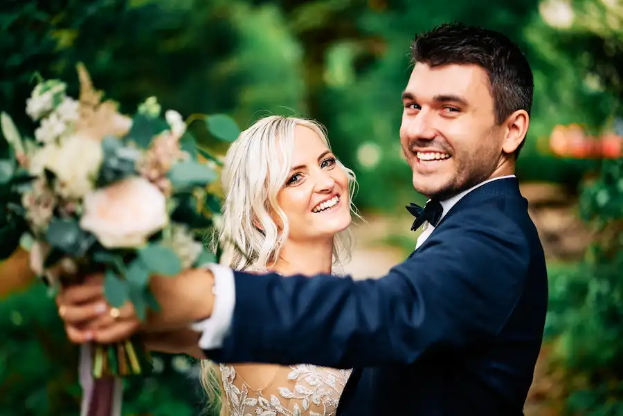 Eine lächelnde Braut und ein lächelnder Bräutigam posieren im Freien, wobei der Bräutigam einen Blumenstrauß in die Kamera hält. Üppiges grünes Laub umgibt sie und fängt den glücklichen Moment ein, als Jessie und Marcel JA sagen - heiraten in Deidesheim. Onelovephoto - Hochzeitsfotografie in Mannheim und Heidelberg