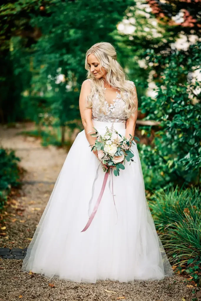 Eine Braut mit langen, gewellten blonden Haaren steht im Freien auf einem Weg in Deidesheim. Sie trägt ein weißes Hochzeitskleid mit Spitzenmieder und Tüllrock, hält Blumen und blickt nach unten - Jessie und Marcel sagen JA, umgeben von grüner Natur. Onelovephoto - Hochzeitsfotografie in Mannheim und Heidelberg