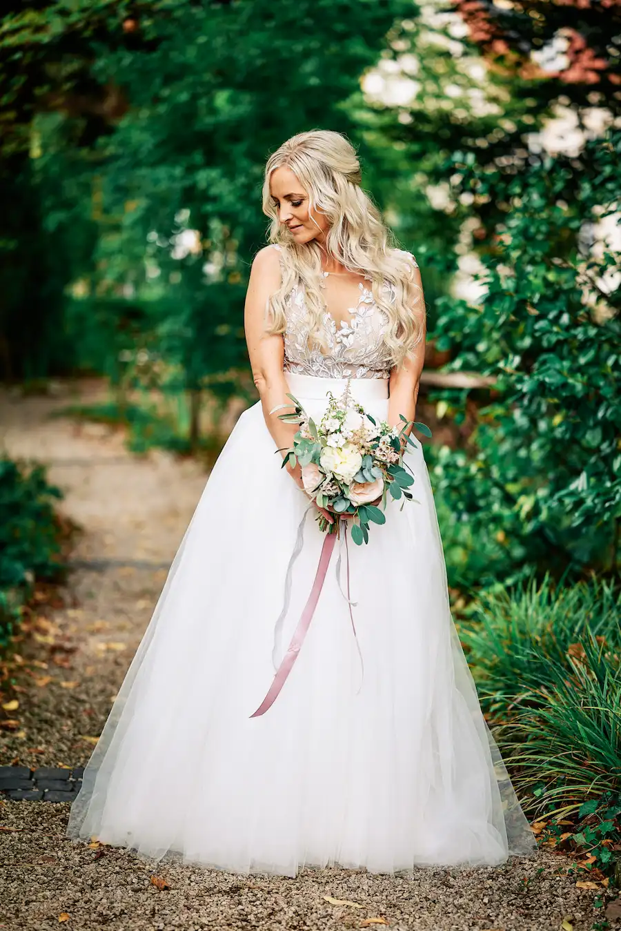 Eine Braut mit langen, gewellten blonden Haaren in einem weißen Spitzenhochzeitskleid steht im Freien, hält einen Blumenstrauß in der Hand, blickt nach unten und lächelt, umgeben von Grünpflanzen - wunderschön eingefangen von Hochzeitsfotograf-Deidesheim.