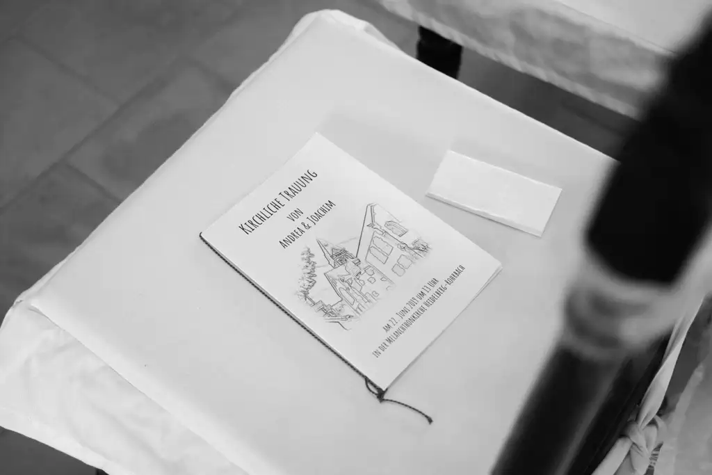 Onelovephoto Ein Schwarz-Weiß-Foto eines Büchleins mit dem Titel "Kirchliche Trauung von Annel & Alexander", mit einer Zeichnung einer Kirche und von Häusern auf dem Umschlag, auf einem weißen Tuch liegend, neben einem kleinen weißen Umschlag. Hochzeitsfotografie in Mannheim und Heidelberg