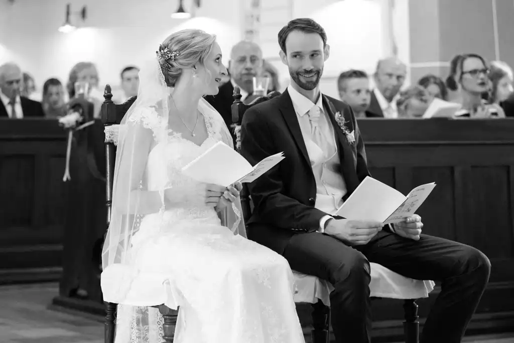 Onelovephoto Braut und Bräutigam sitzen während ihrer Hochzeitszeremonie lächelnd nebeneinander und halten ein Programm in der Hand, während die Gäste hinter ihnen Platz nehmen. Das Bild ist in schwarz-weiß gehalten. Hochzeitsfotografie in Mannheim und Heidelberg