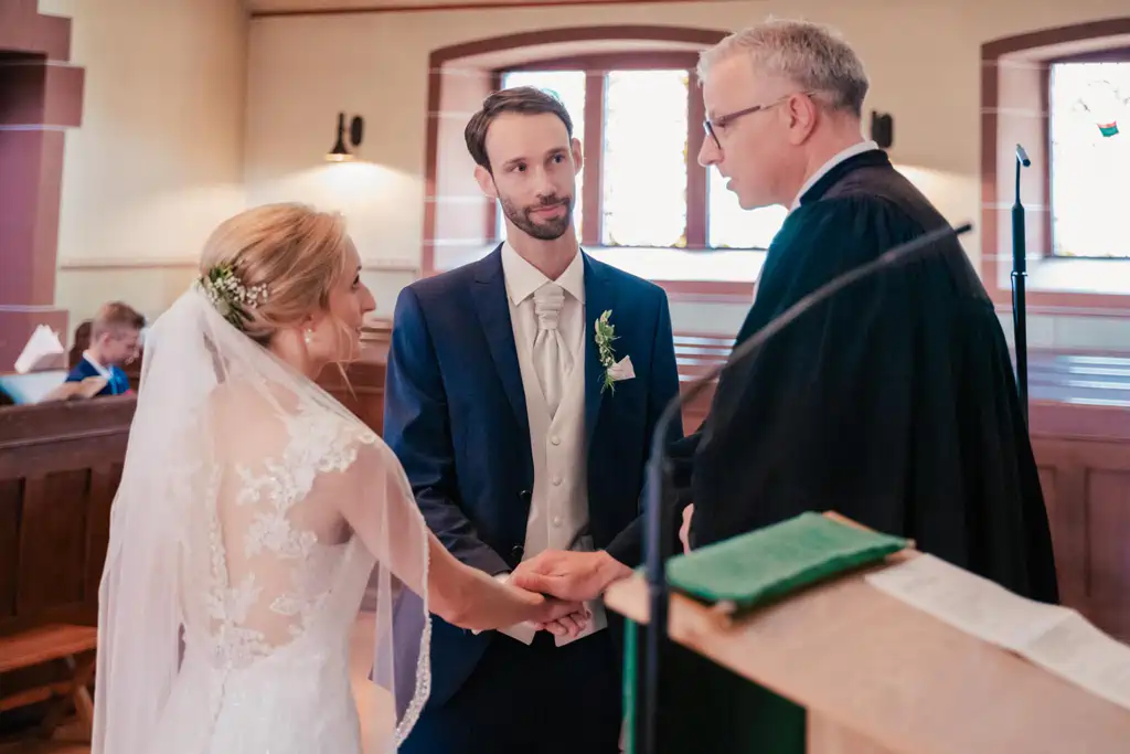 Onelovephoto Braut und Bräutigam stehen während ihrer Trauung in einer Kirche Hand in Hand vor dem Pfarrer, während das natürliche Licht durch die Fenster hinter ihnen fällt. Hochzeitsfotografie in Mannheim und Heidelberg