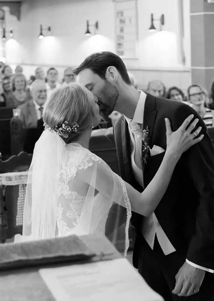 Braut und Bräutigam küssen sich während ihrer Hochzeitszeremonie im Hotel Grenzhof vor dem Altar, während die Gäste im Hintergrund lächeln. Das Schwarz-Weiß-Foto wurde von einem Hochzeitsfotografen aufgenommen und hebt ihren geblümten Schleier und seinen Anzug hervor Braut und Bräutigam küssen sich während ihrer Hochzeitszeremonie im Hotel Grenzhof vor dem Altar, während die Gäste im Hintergrund lächeln. Das Schwarz-Weiß-Foto wurde von einem Hochzeitsfotografen aufgenommen und hebt ihren geblümten Schleier und seinen Anzug hervor.