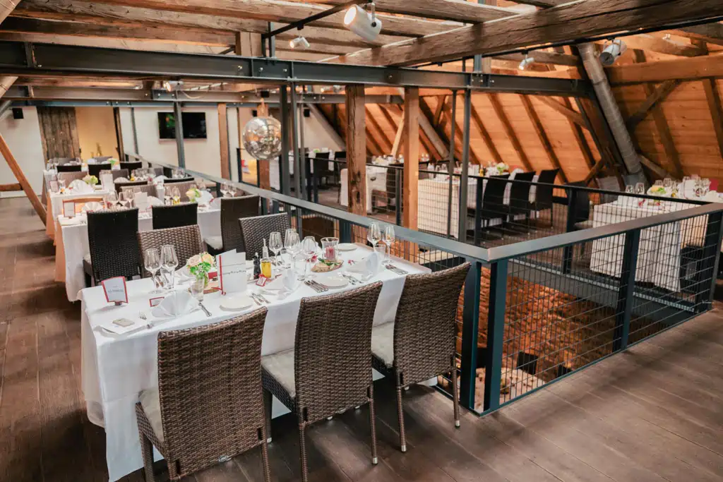 Onelovephoto Ein modernes, elegantes Restaurant mit ordentlich gedeckten Tischen, weißen Tischtüchern, Korbstühlen und Weingläsern. Der Raum verfügt über Holzbalken, warme Beleuchtung und einen oberen Mezzanin-Essbereich. Hochzeitsfotografie in Mannheim und Heidelberg