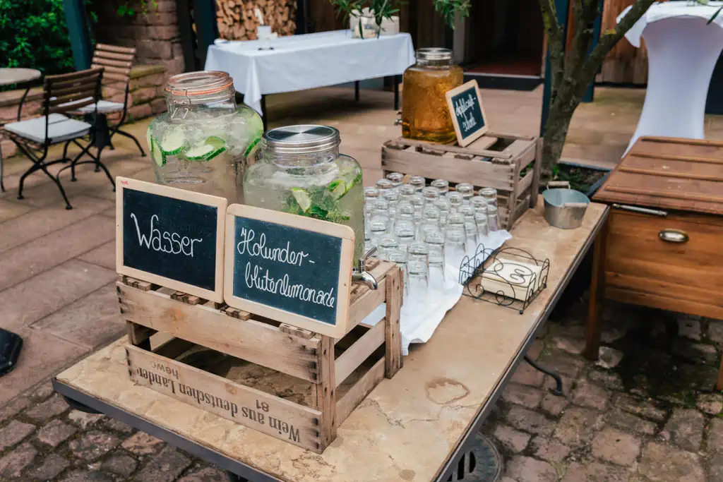 Onelovephoto Eine Getränkestation im Freien mit großen Gläsern mit Wasser und Holunderblütenlimonade, beschriftet mit Kreideschildern, neben Reihen von leeren Gläsern auf einem Holztisch. Hochzeitsfotografie in Mannheim und Heidelberg