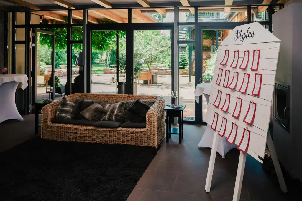 Onelovephoto Ein gemütlicher Innenraum mit einem Korbsofa und dunklen Kissen befindet sich neben einem großen Fenster. Ein weißer Holzsitzplan mit der Aufschrift "Sitzplan" und rot umrandeten Karten steht im Vordergrund. Hochzeitsfotografie in Mannheim und Heidelberg