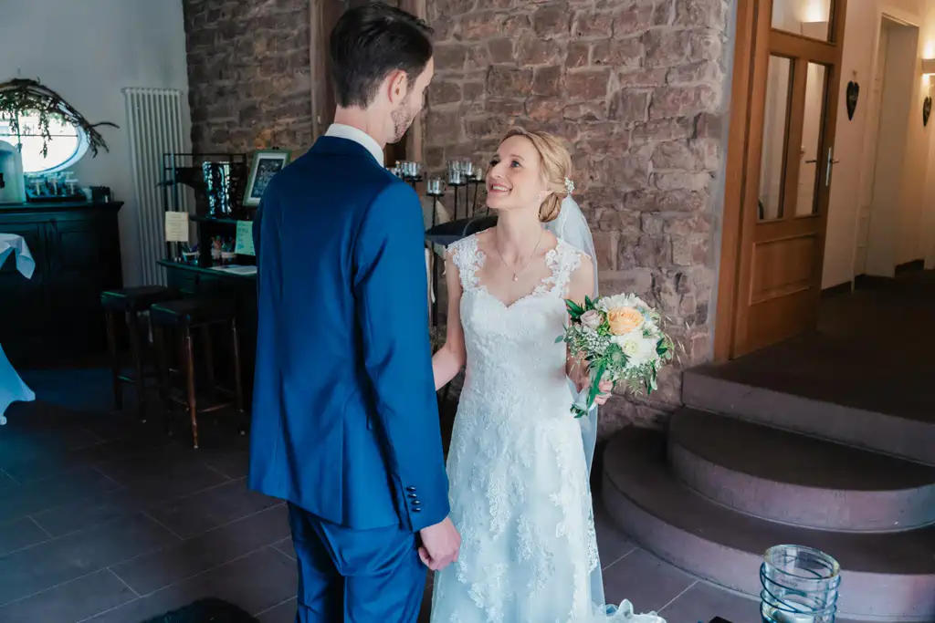 Onelovephoto Eine Braut in einem weißen Spitzenkleid mit einem Blumenstrauß in der Hand lächelt einem Bräutigam in einem blauen Anzug in einem warm beleuchteten Raum mit Steinwänden und Holzmöbeln zu. Hochzeitsfotografie in Mannheim und Heidelberg