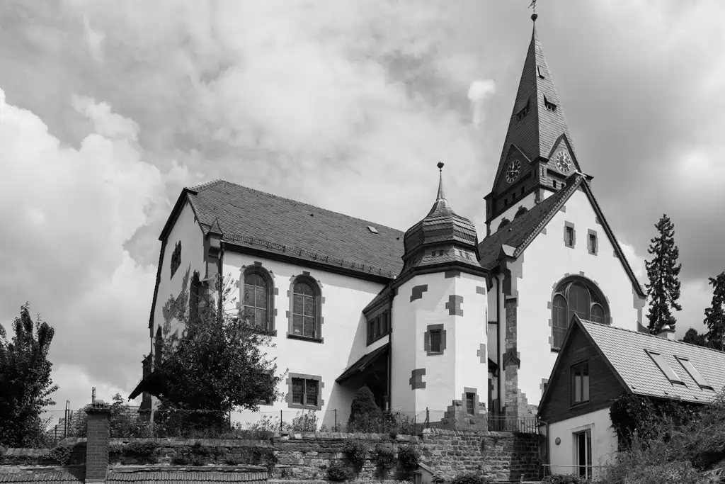 Onelovephoto Schwarz-Weiß-Foto einer historischen Kirche mit gewölbten Fenstern, einem hohen Kirchturm mit Uhr und einem kleinen Nebengebäude vor einem bewölkten Himmel, umgeben von einer Steinmauer und Laub. Hochzeitsfotografie in Mannheim und Heidelberg