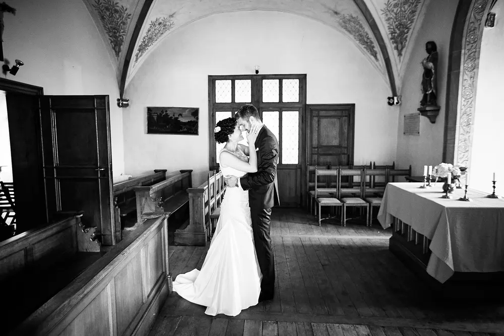 Braut und Bräutigam umarmen und küssen sich innig in einer wunderschön dekorierten, leeren Kapelle mit verzierten Decken und Holzbänken - perfekt für Heiraten auf der Mittelburg in Neckarsteinach. Die Szene ist in Schwarz-Weiß aufgenommen. Onelovephoto - Hochzeitsfotografie in Mannheim und Heidelberg