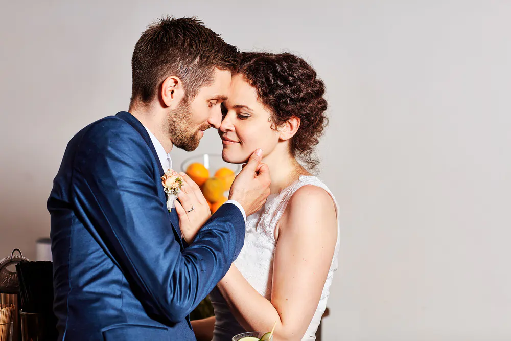 Onelovephoto Ein Bräutigam in einem blauen Anzug hält zärtlich das Kinn einer Braut in einem weißen Kleid, während sie sich eng aneinander lehnen und ihre Stirnen vor einem einfachen Hintergrund sanft lächelnd berühren. Hochzeitsfotografie in Mannheim und Heidelberg