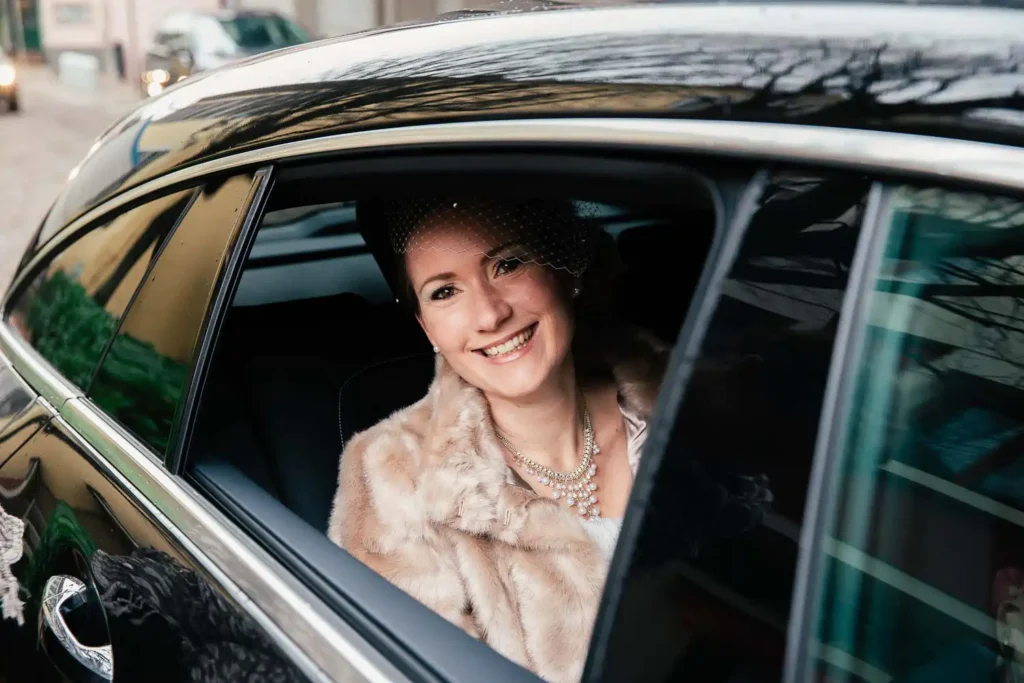 Onelovephoto Eine lächelnde Frau in Pelzmantel und Schleier sitzt auf dem Rücksitz eines Autos und schaut aus dem offenen Fenster, in dem sich Gebäude und Bäume auf der glänzenden Oberfläche des Autos spiegeln. Hochzeitsfotografie in Mannheim und Heidelberg