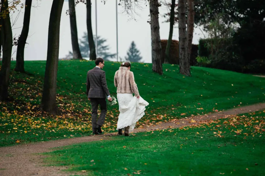 Onelovephoto Eine Braut und ein Bräutigam gehen in einem grünen Park einen mit Blättern übersäten Weg entlang, wobei die Braut ihr Kleid und der Bräutigam einen Blumenstrauß hält, umgeben von hohen Bäumen. Hochzeitsfotografie in Mannheim und Heidelberg