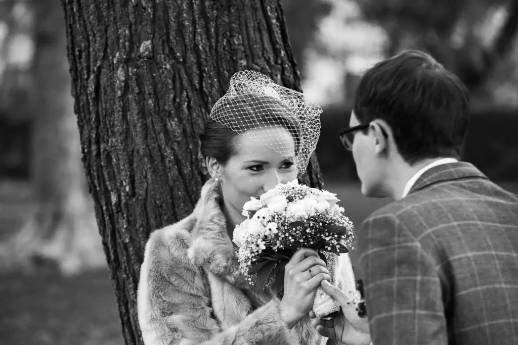 Onelovephoto Eine Frau mit Pelzmantel und Vogelkäfigschleier hält einen Blumenstrauß und lächelt einen Mann mit Brille und kariertem Anzug an. Sie stehen dicht beieinander im Freien in der Nähe eines Baumes. Das Bild ist in schwarz-weiß gehalten. Hochzeitsfotografie in Mannheim und Heidelberg