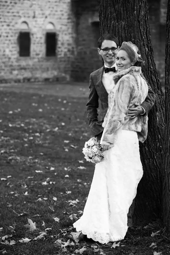 Onelovephoto Eine Braut in einem Spitzenkleid und einer Pelzstola steht an einem Baum und hält einen Blumenstrauß in der Hand, während der Bräutigam in Anzug und Fliege hinter ihr steht. Beide lächeln draußen in der Nähe eines steinernen Gebäudes auf einem mit Blättern übersäten Rasen. Hochzeitsfotografie in Mannheim und Heidelberg