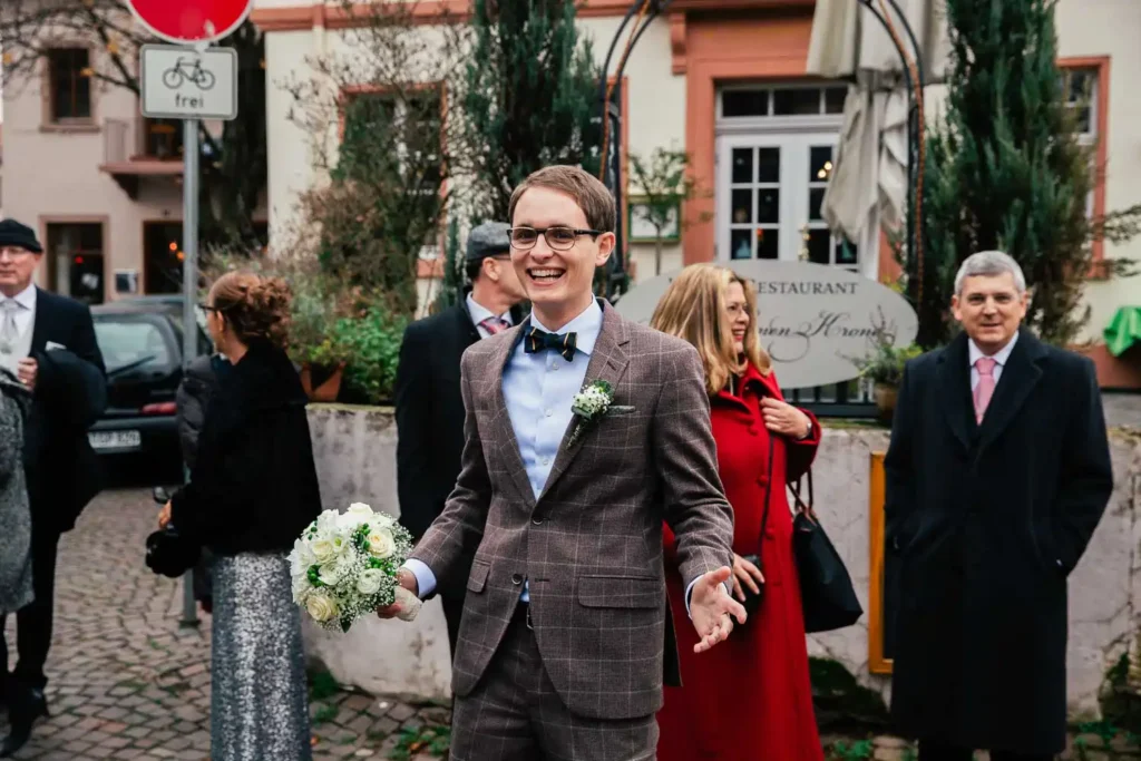 Onelovephoto Ein lächelnder Mann in einem karierten Anzug mit einem Blumenstrauß steht im Freien, umgeben von gut gekleideten Menschen. Der Hintergrund zeigt einen Restauranteingang, Bäume und Kopfsteinpflaster, was auf eine feierliche Veranstaltung hindeutet. Hochzeitsfotografie in Mannheim und Heidelberg