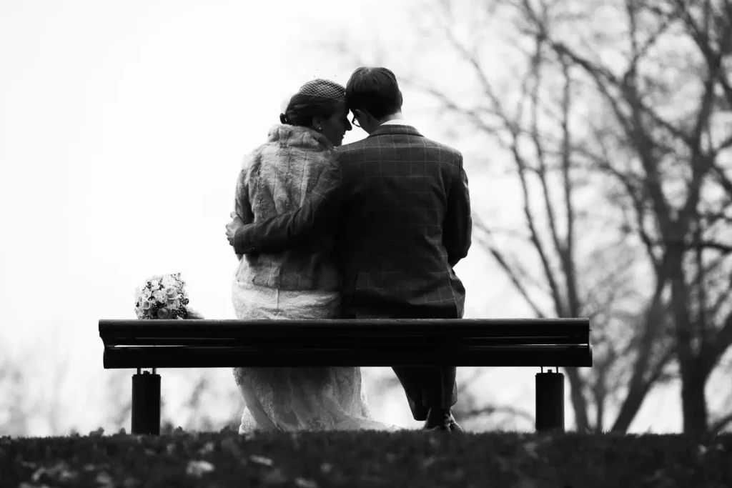 Onelovephoto Ein Paar in Hochzeitskleidung sitzt eng beieinander auf einer Parkbank, umarmt sich und berührt sich an der Stirn. Im Hintergrund sind kahle Bäume zu sehen und ein Blumenstrauß liegt neben ihnen. Das Bild ist in schwarz-weiß gehalten. Hochzeitsfotografie in Mannheim und Heidelberg