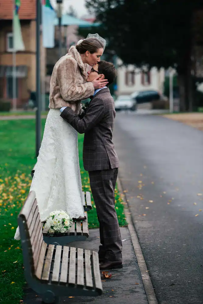 Onelovephoto Eine Braut in einem weißen Kleid und einer Kunstpelzjacke steht auf einer Parkbank und küsst einen Bräutigam in einem karierten Anzug, der auf dem Boden steht und ihre Taille hält. Ein Blumenstrauß liegt auf der Bank neben den beiden. Hochzeitsfotografie in Mannheim und Heidelberg