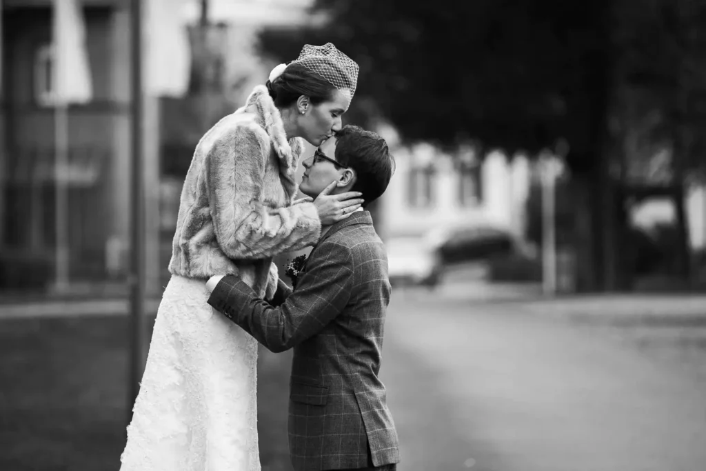 Als Hochzeitsfotograf in Ladenburg fange ich intime Momente ein: Eine Braut im Pelzstola und Schleier küsst den knienden Bräutigam auf der Stirn, während er sie liebevoll hält - eine zeitlose Szene auf einer ruhigen Straße in Schwarz-Weiß. Onelovephoto - Hochzeitsfotografie in Mannheim und Heidelberg