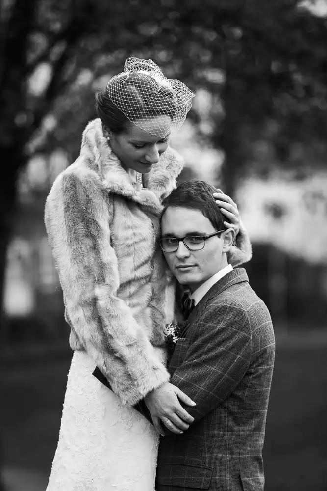 Onelovephoto Ein Schwarz-Weiß-Foto eines Paares im Freien; die Frau in Pelzmantel und Vogelkäfigschleier umarmt den Mann mit Brille und kariertem Anzug, der in die Kamera schaut, während sie liebevoll auf ihn herabblickt. Hochzeitsfotografie in Mannheim und Heidelberg