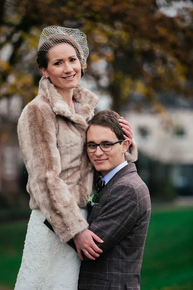 Eine lächelnde Braut in einem Spitzenkleid und einer Pelzjacke umarmt den Bräutigam, Janina und Florian, mit Brille und einem karierten Anzug im Freien, mit verschwommenen Bäumen und Gebäuden im Hintergrund. Aufgenommen von Hochzeitsfotograf Ladenburg.