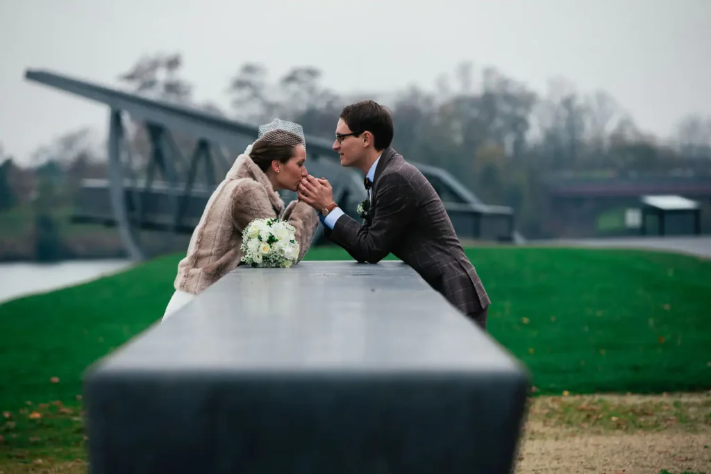 Als Hochzeitsfotograf in Ladenburg fange ich besondere Momente ein: Ein Brautpaar lehnt sich auf einer Betonfläche im Freien aneinander, hält Händchen und blickt sich verliebt in die Augen - vor grüner Kulisse an einem bewölkten Tag. Onelovephoto - Hochzeitsfotografie in Mannheim und Heidelberg