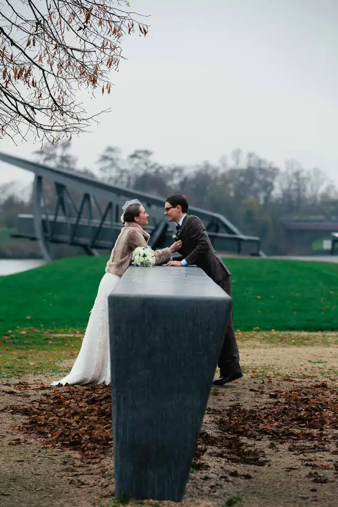 Onelovephoto Eine Braut und ein Bräutigam stehen sich an einem langen Steintisch im Freien gegenüber, halten sich an den Händen und lächeln sich an, mit einer modernen Brücke und grünem Gras im Hintergrund. Hochzeitsfotografie in Mannheim und Heidelberg